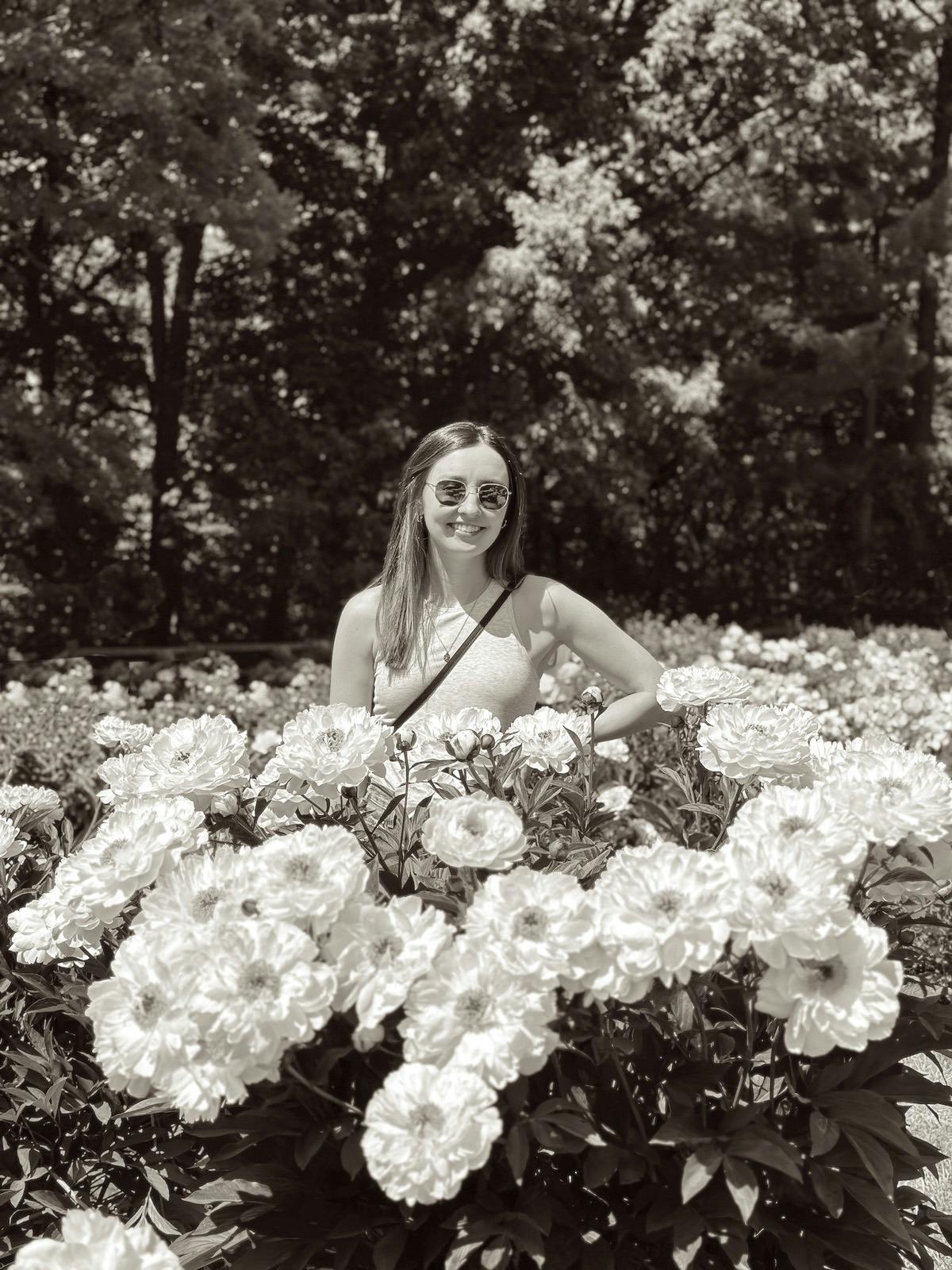 Black and white photo of Jennifer wearing sunglasses, smiling during a sunny day in a garden full of peonies